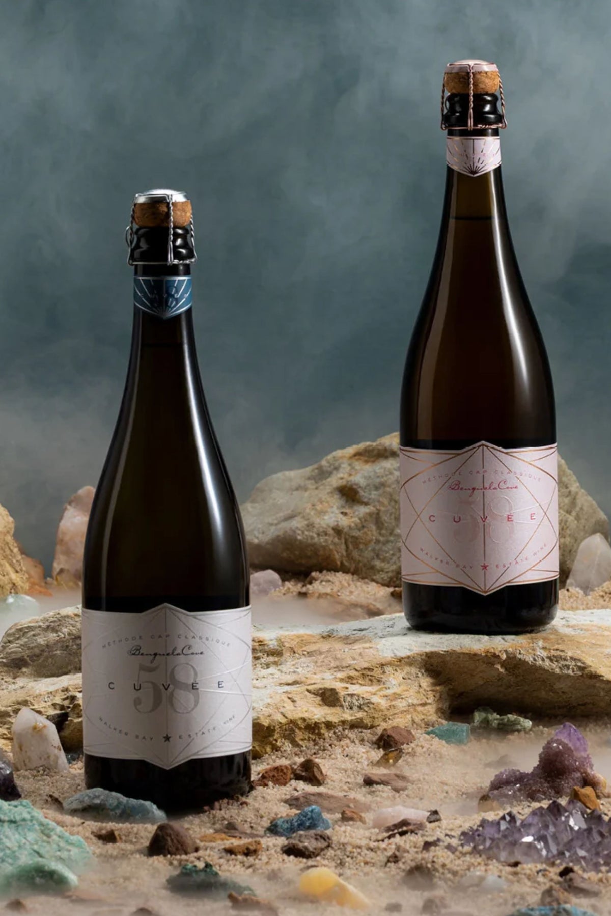 Two dark glass bottles of Cuvier sparkling wine with white labels, styled on a sandy surface with rocks and colorful crystals against a smoky blue-gray background.
